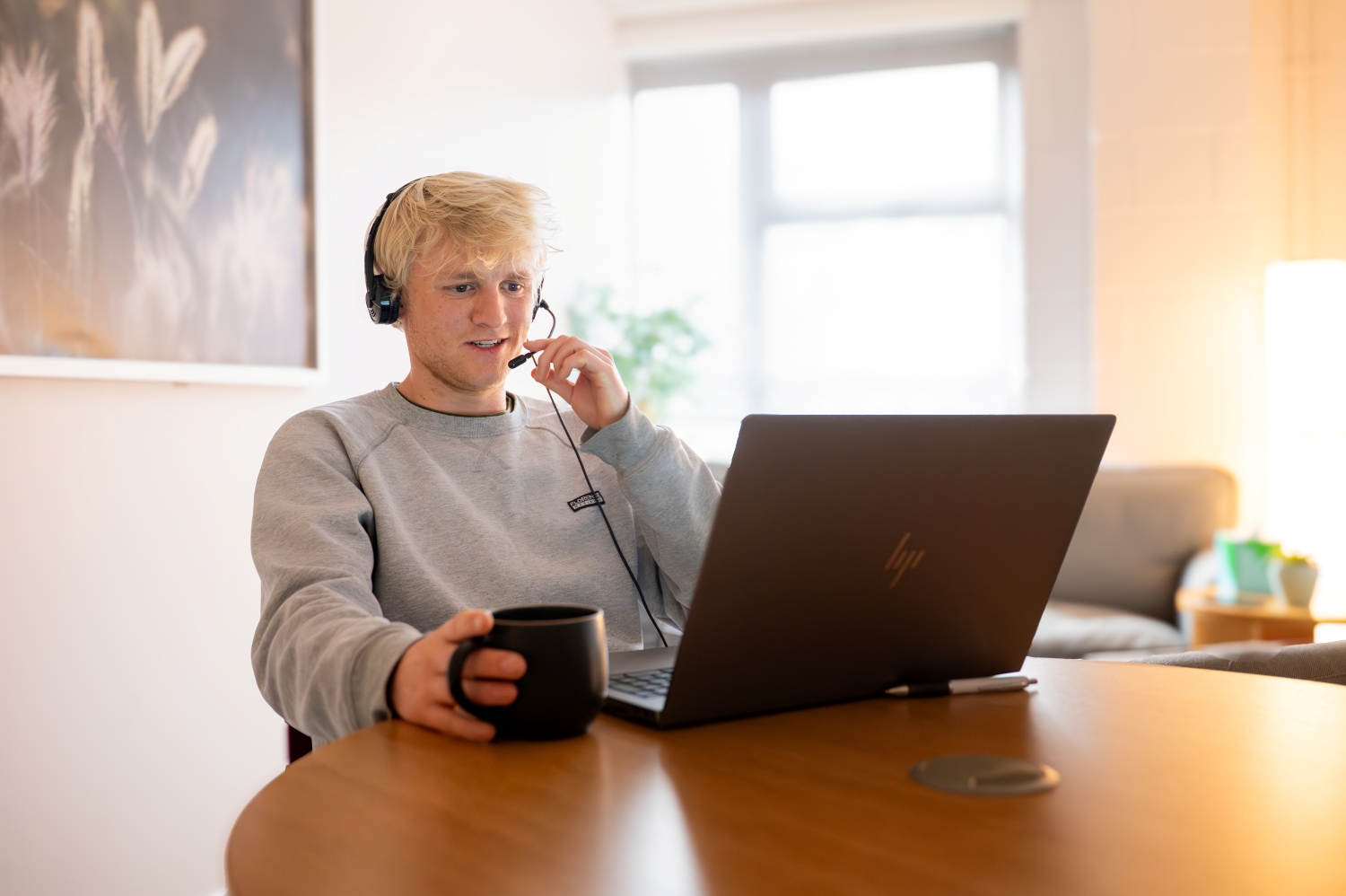 Student studying online from home, wearing a headset and using a laptop with a coffee mug in hand, in a bright, relaxed living space