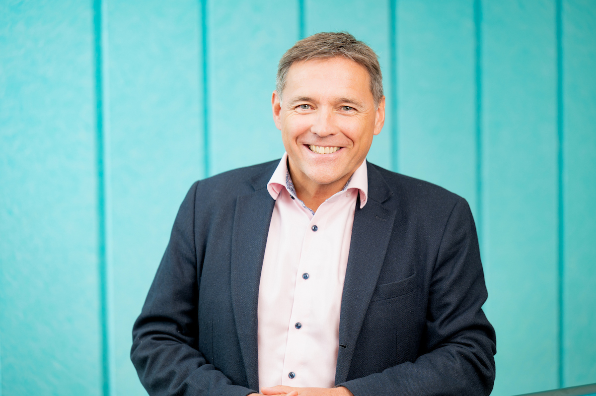 Derek Bond smiling at the camera with a teal blue background Derek Bond smiling at the camera with a teal blue background