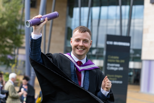 George Gunn in his graduation gown, holding up his scroll and smiling