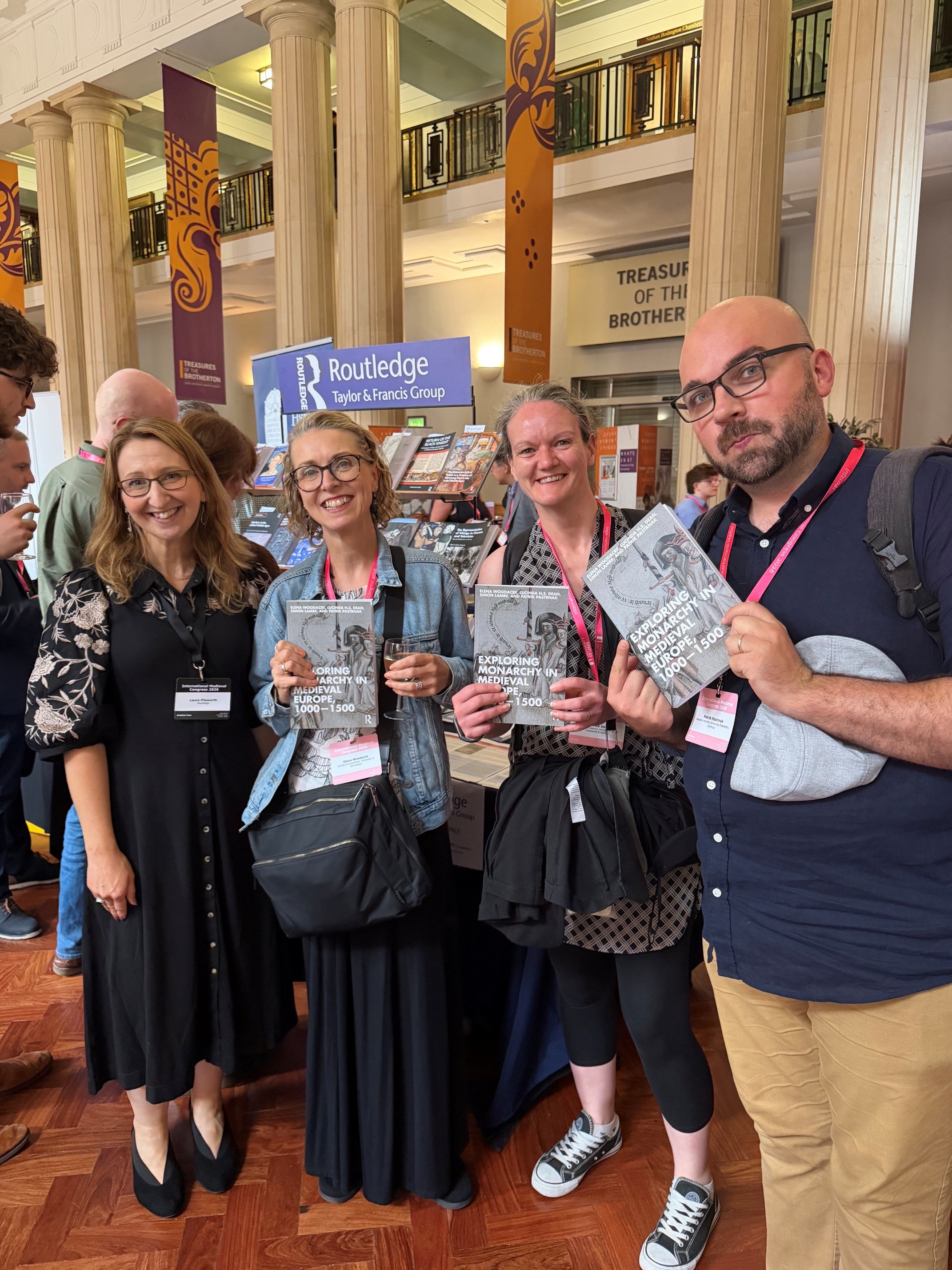 Four people at a publishing event, holding copies of Exploring Monarchy in Medieval Europe, 1000-1500, with a Routledge display in the background Four people at a publishing event, holding copies of Exploring Monarchy in Medieval Europe, 1000-1500, with a Routledge display in the background