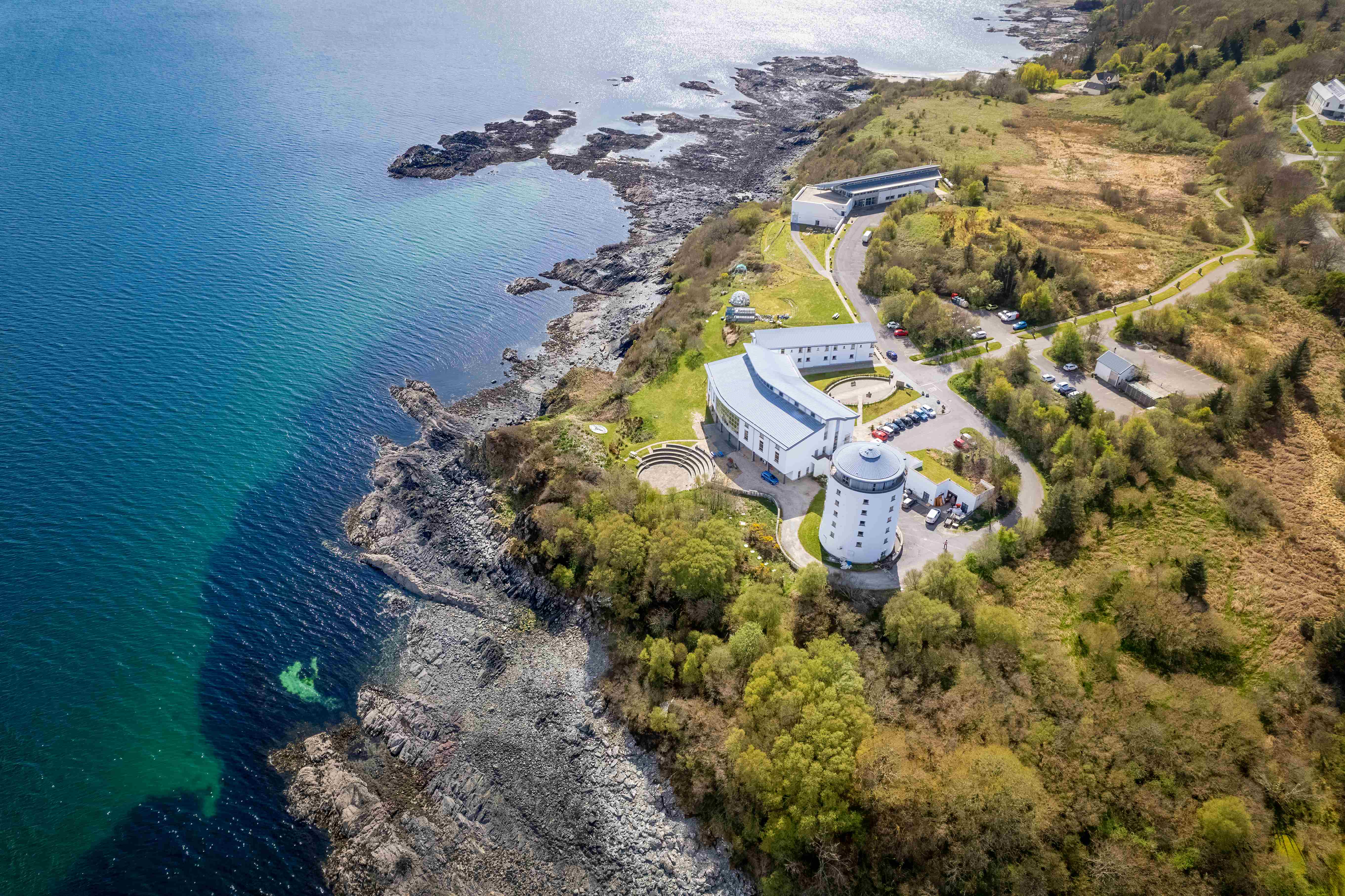 Aerial photo of a white building set by a rocky seashore, surrounded by greenery.