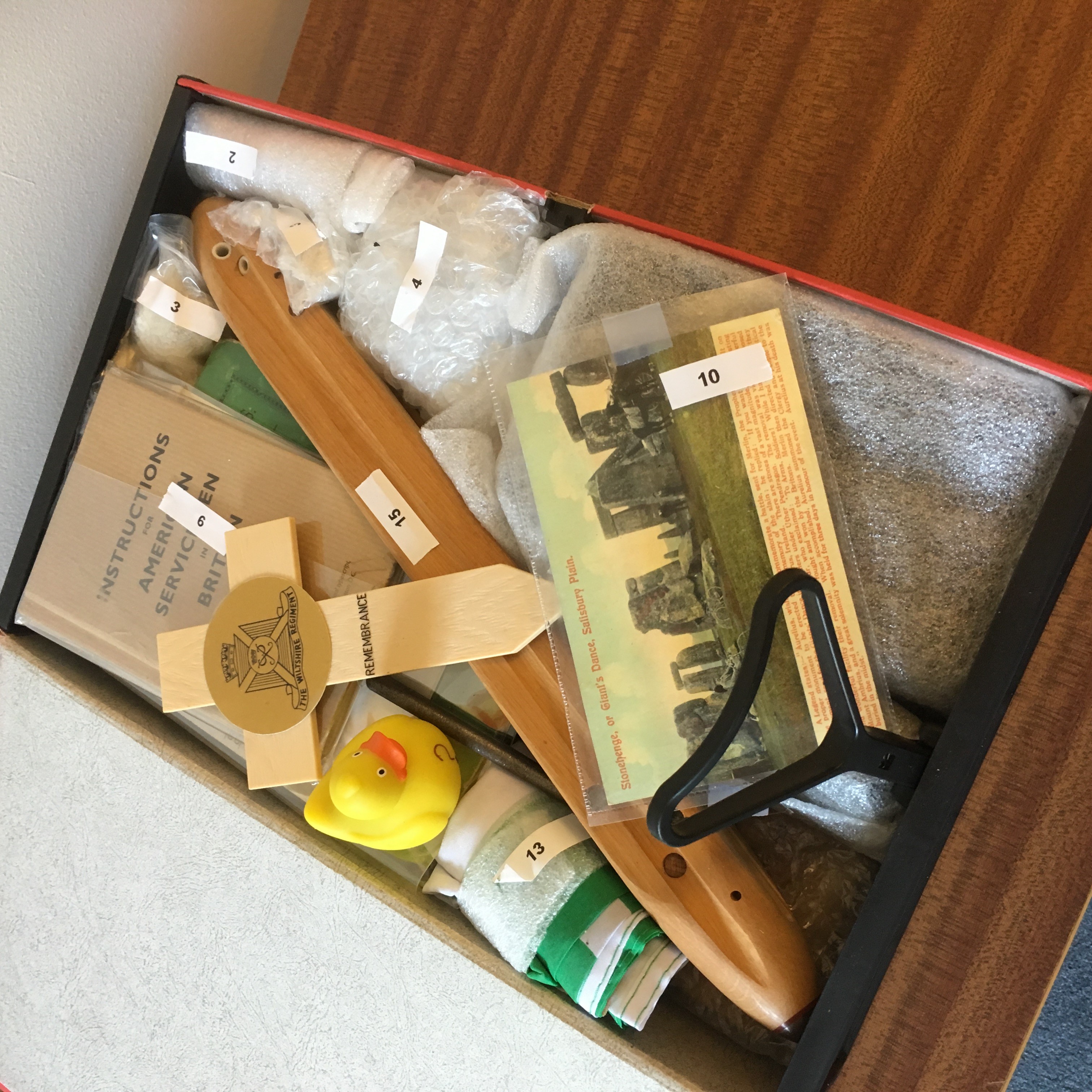 A collection of items packed in a red box, including a small wooden remembrance cross, a yellow rubber duck, postcard of the Stonehenge and a book titled Instructions to American Servicewomen in Britain among other things. Items are numbered with labels.