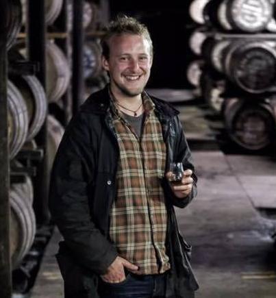 Man, standing among whisky barrels, holds a glencairn glass with a dram