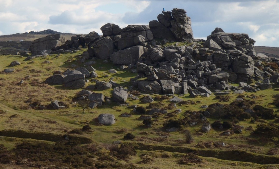 Landscape at Dartmoor with a large formation of rocks taking the centre stage and a lone climber sitting on top
