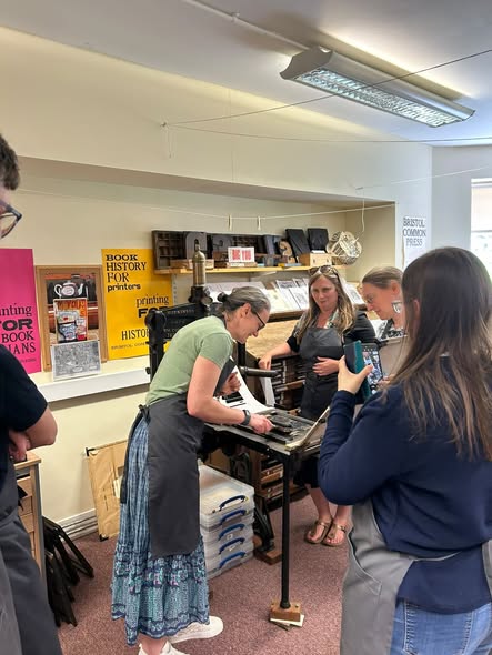 Woman wearing an apron bending over a manual printing press. Group of four people observe them working, and posters on the walls and prints on the shelves showcase different kinds of printing products Woman wearing an apron bending over a manual printing press. Group of four people observe them working, and posters on the walls and prints on the shelves showcase different kinds of printing products