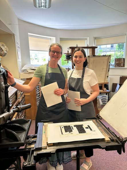 Two women wearing aprons standing next to a manual printing press Two women wearing aprons standing next to a manual printing press