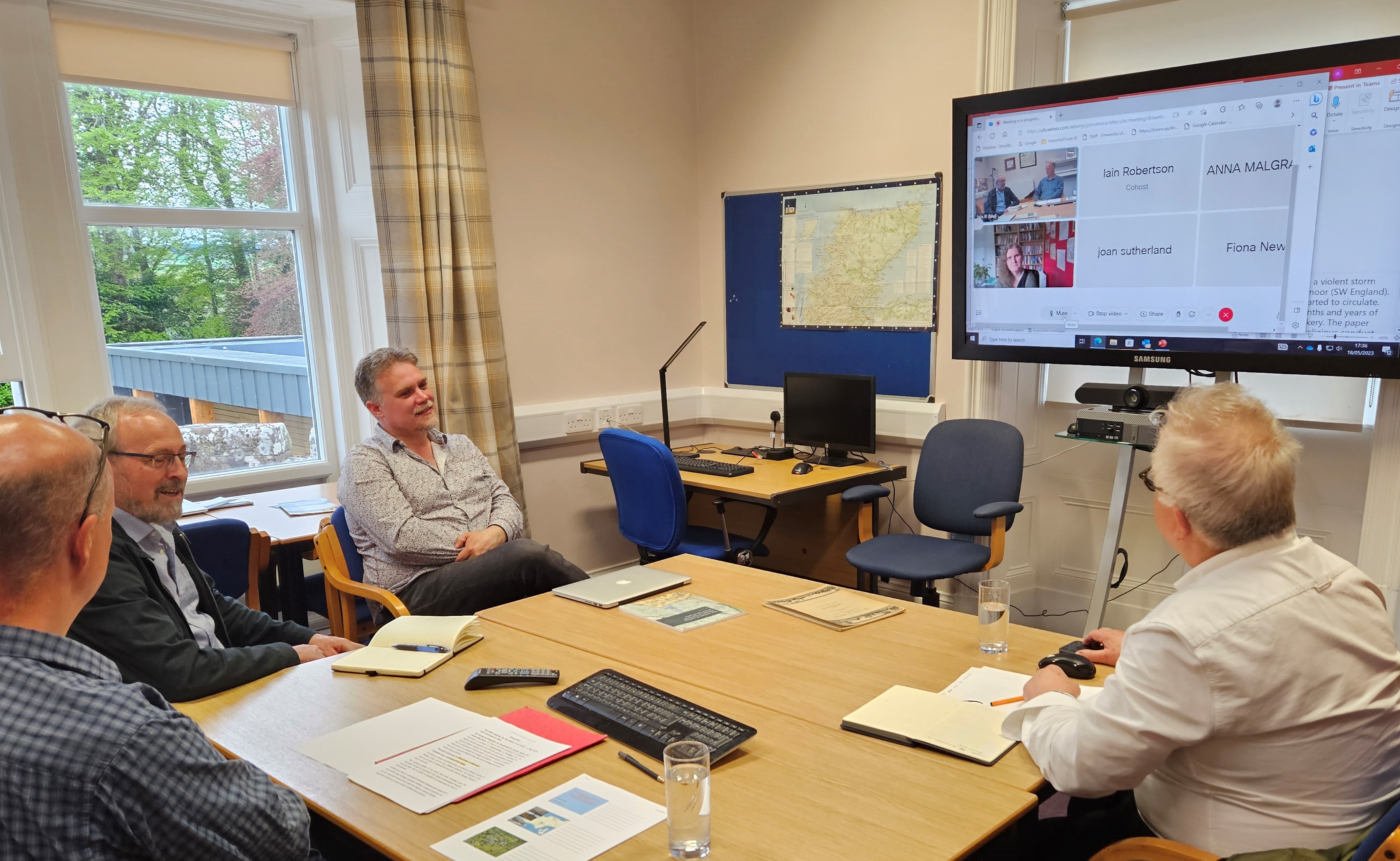 Four men seated around a table looking at a large screen with details of online attendees shown on it. Four men seated around a table looking at a large screen with details of online attendees shown on it.