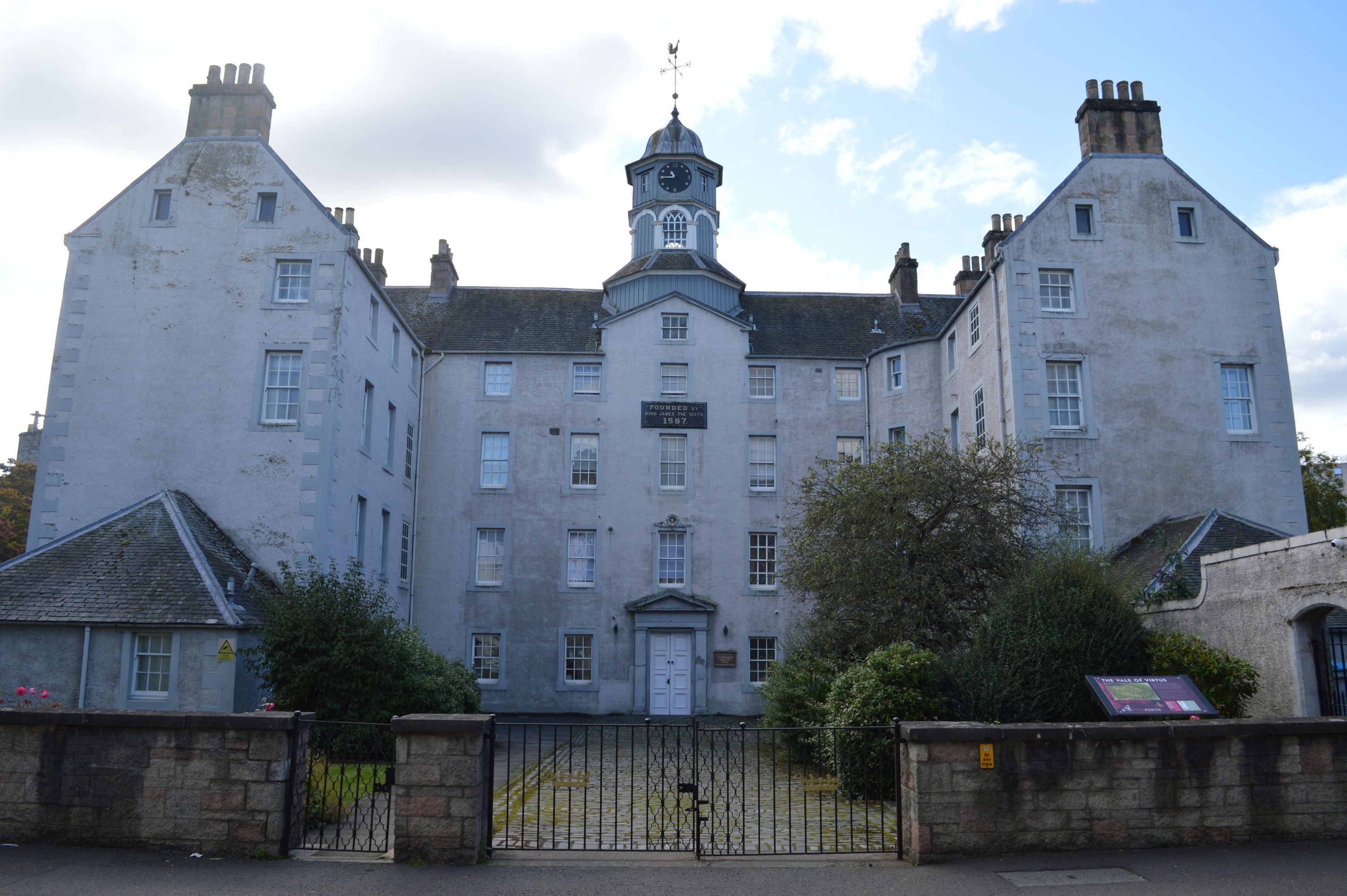 Grey-coloured building with two arms protruding towards the camera. Stonewall and gate in front of the building are framed by leafy shrubs and trees.