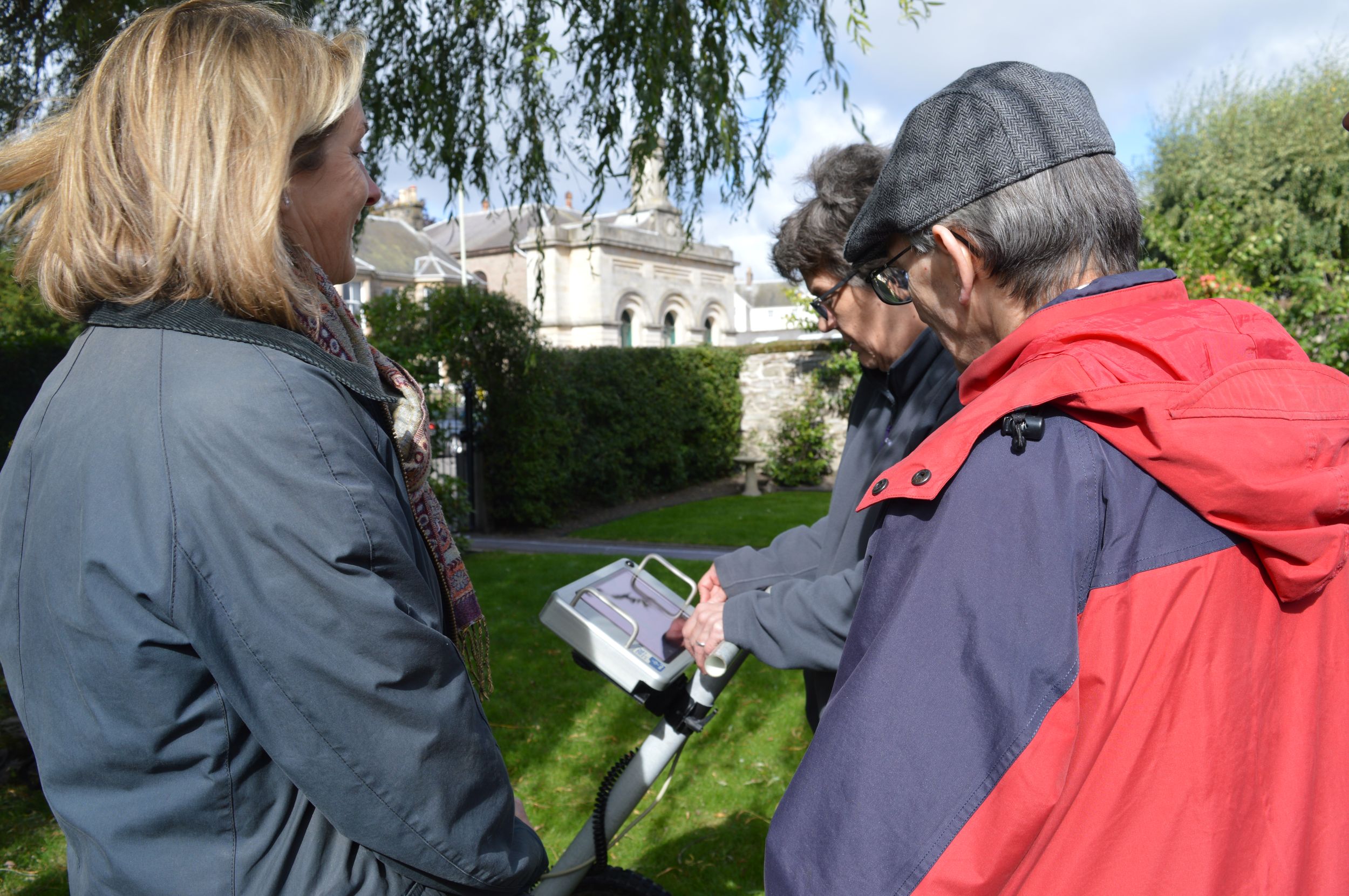 Three people gathered around of a screen on a ground penetrating radar scanner. In the background there are stone buildings and leafy trees.