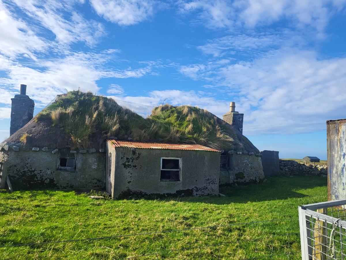 A stone house with a thatched roof that now has grass growing from it. 