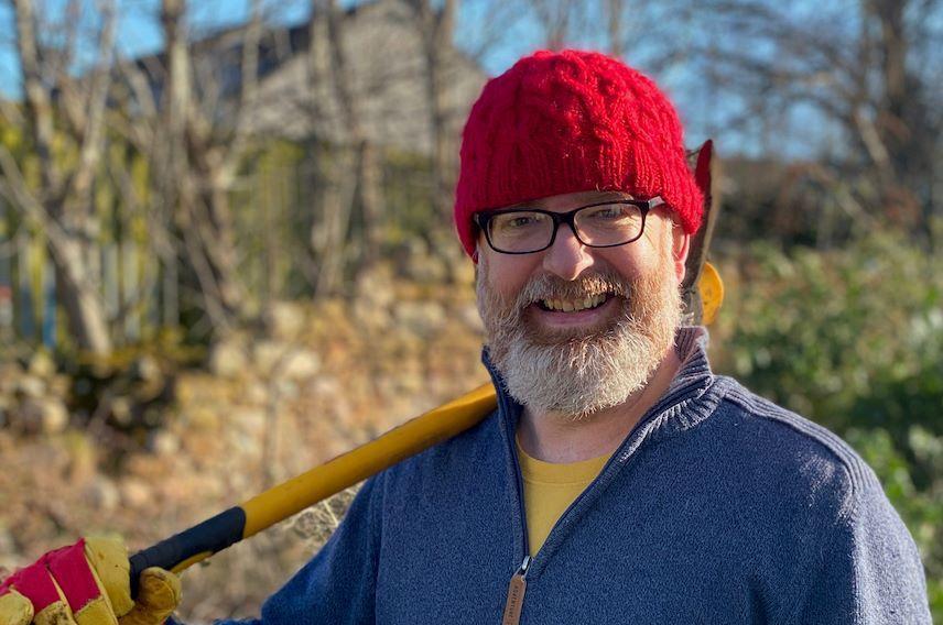 Dr Jim MacPherson smiling at the camera while holding a garden tool and wearing a red beanie hat