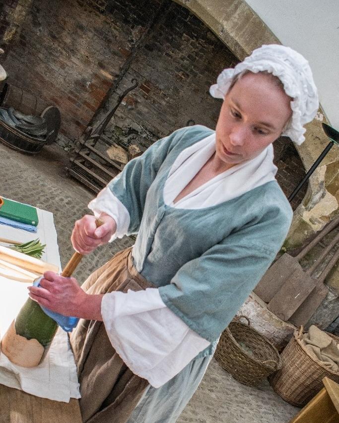 Woman dressed in 18th-century clothing, including an apron and bonnet, churning butter in a setting of historical kitchen utensils and equipment
