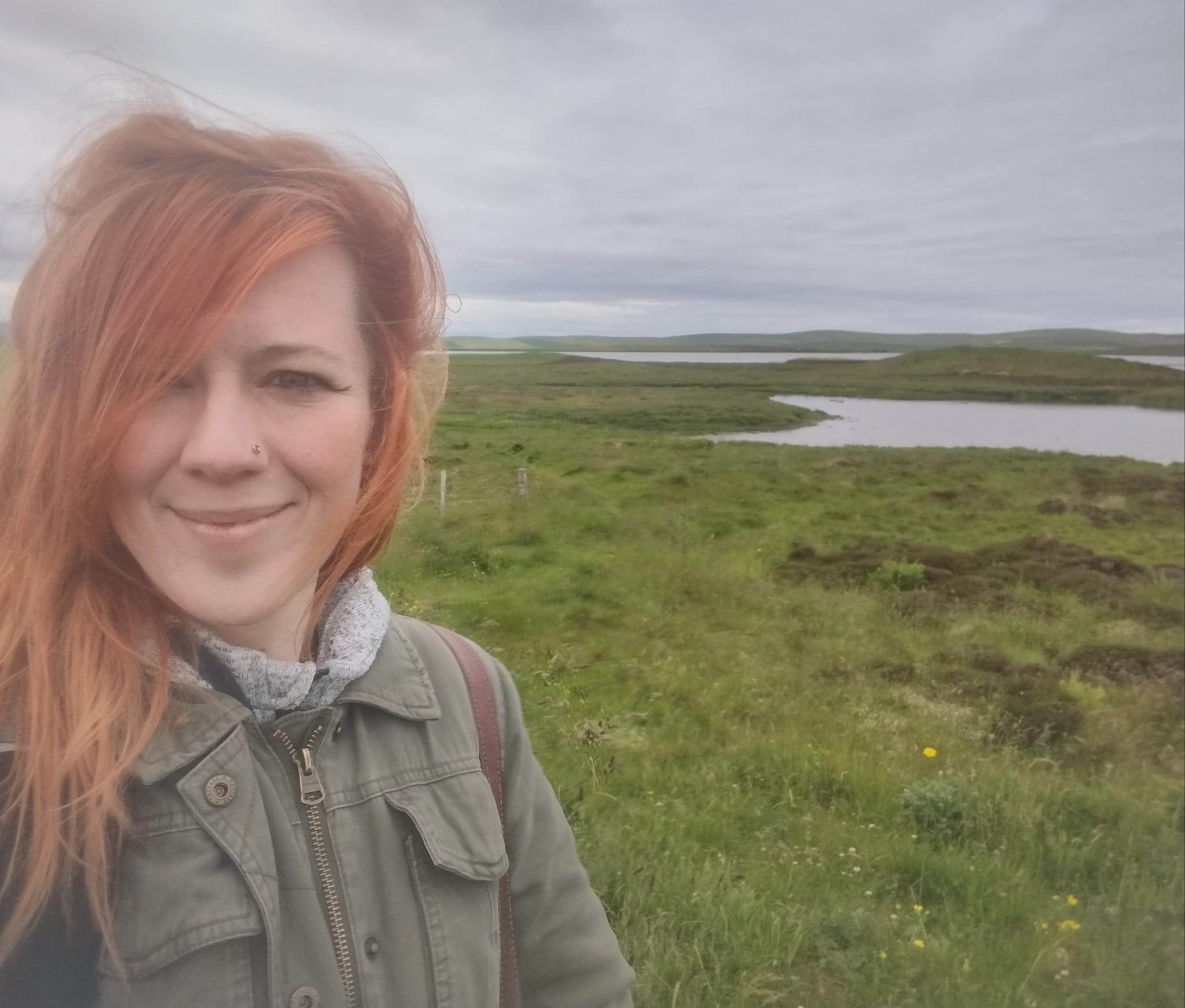 Woman with red hair standing close to the camera with a green moorland landscape behind her, with bodies of water in the background and a grey sky above.