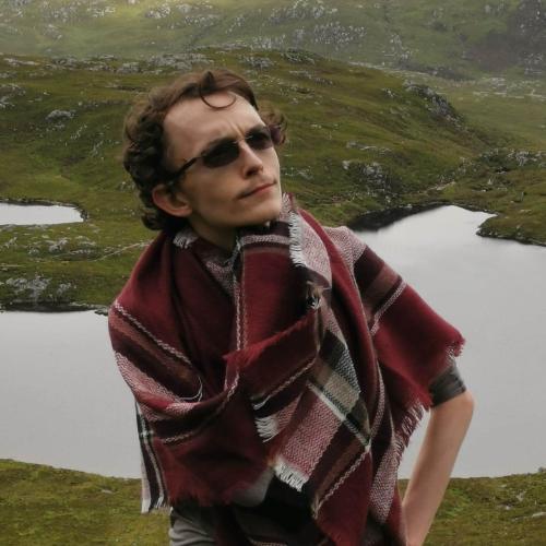 Man wearing sunglasses and scarf standing in the forefront while the background shows a green landscape and a loch
