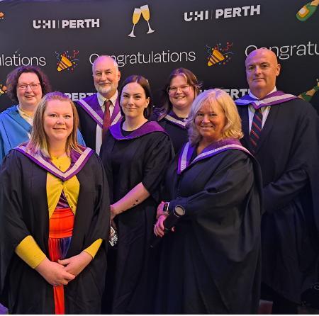 Group of people wearing formal wear and graduations gowns and hoods. Behind them, a backdrop shows repeated texts of UHI Perth and Congratulations, with emojis of champagne flutes and confetti cones