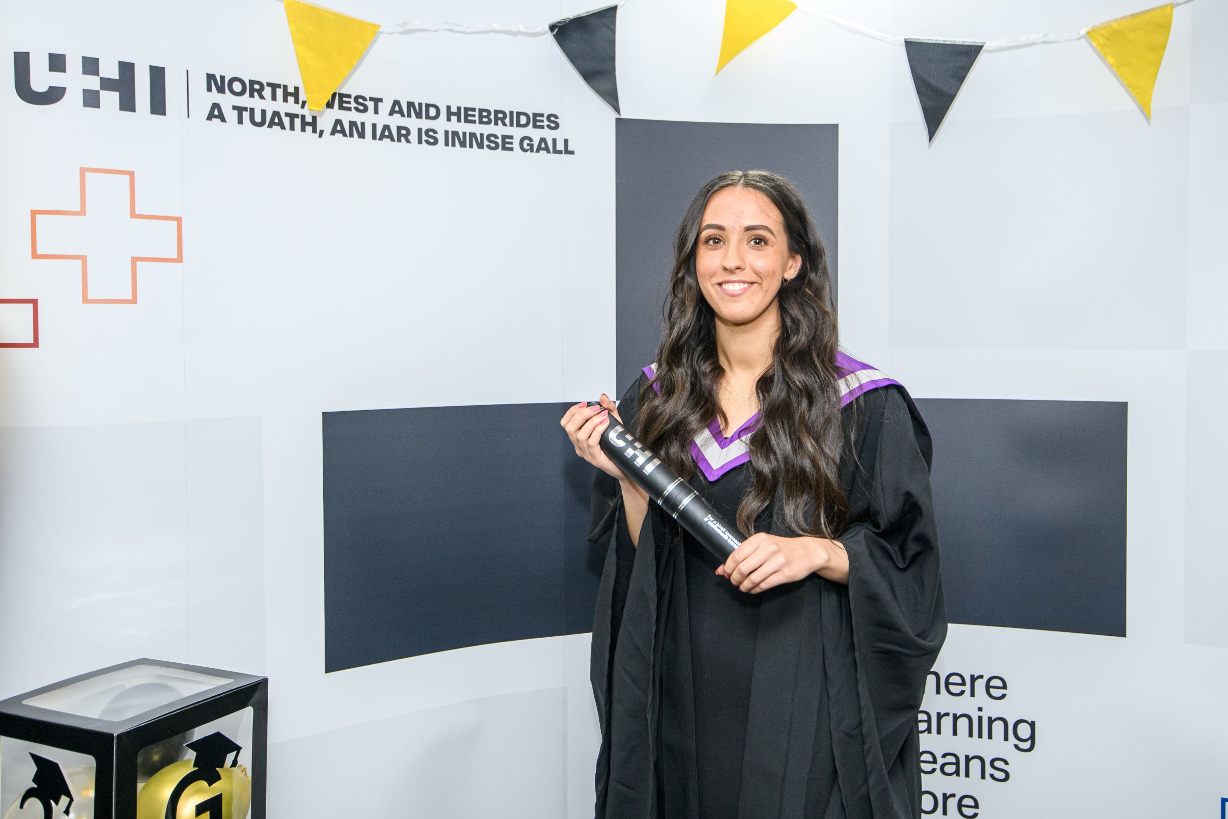 Woman with long dark hair wearing a black dress, a graduation robe and hood, holding a degree scroll. Above her, there is gold and black bunting. Woman with long dark hair wearing a black dress, a graduation robe and hood, holding a degree scroll. Above her, there is gold and black bunting.