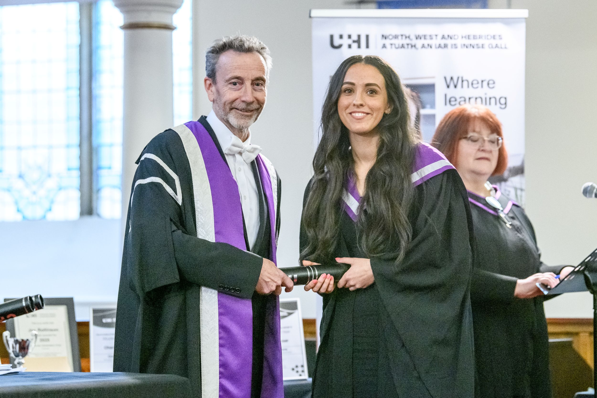 Woman and man in academic gowns and hoods both holding on to a graduation scroll and looking at the camera smiling Woman and man in academic gowns and hoods both holding on to a graduation scroll and looking at the camera smiling