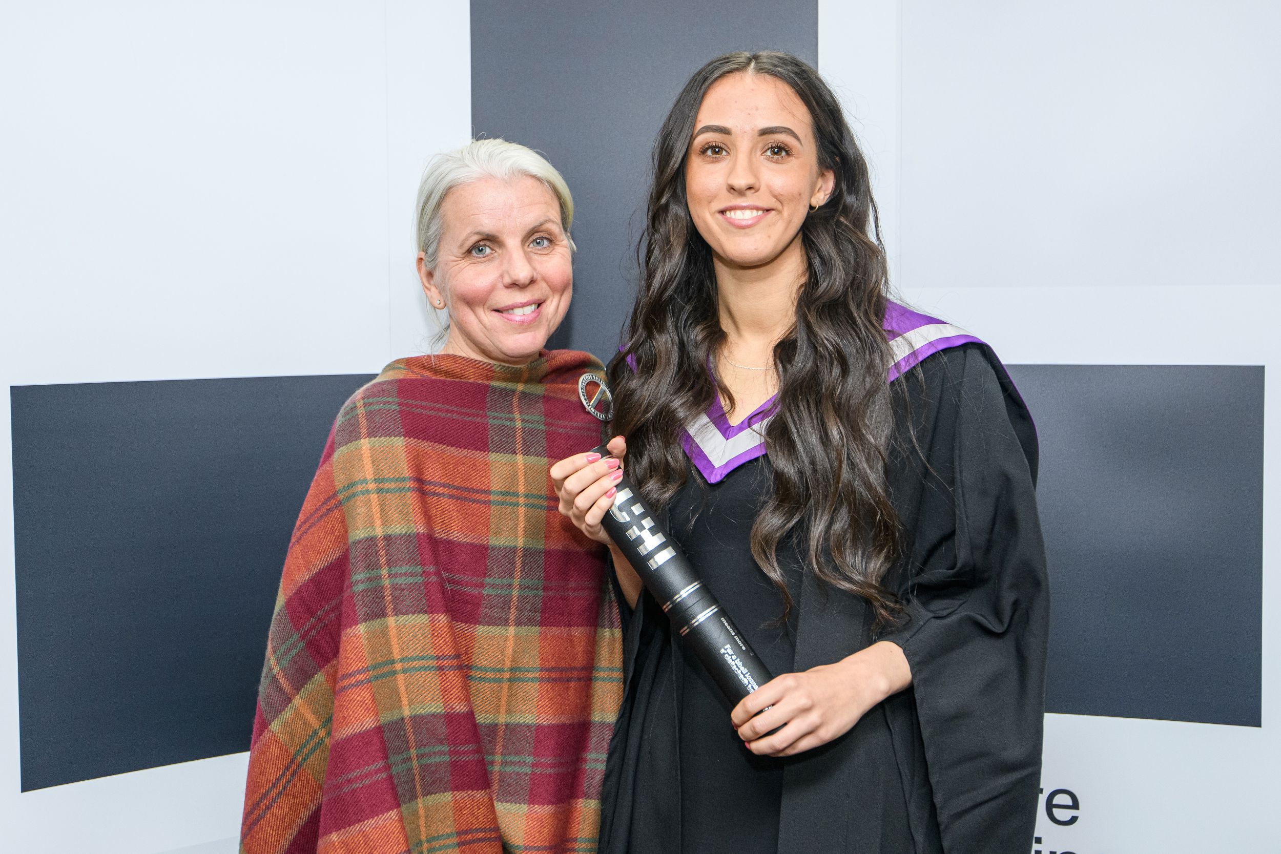 Two women smiling at the camera. The other one is wearing a graduation gown and hood, while the other wears a tartan-patterned shawl. Two women smiling at the camera. The other one is wearing a graduation gown and hood, while the other wears a tartan-patterned shawl.