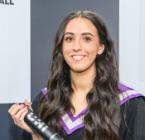 A smiling woman wearing a graduation gown and hood, holding a degree scroll A smiling woman wearing a graduation gown and hood, holding a degree scroll