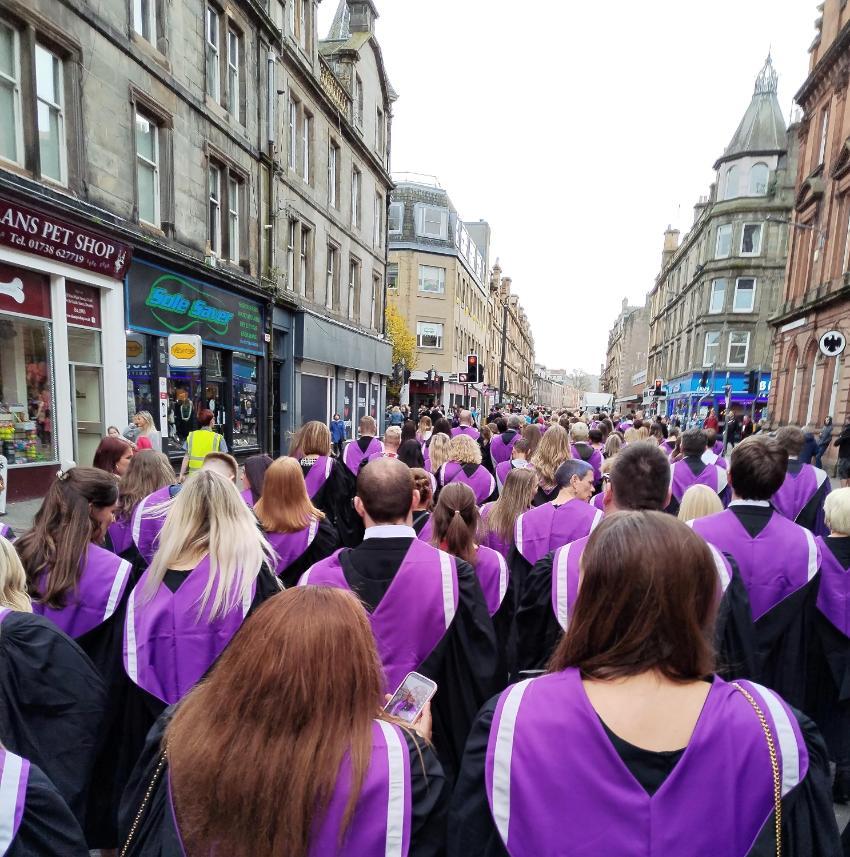 Group of UHI graduates clad in black gowns and purple hoods walking along a street with shop signs and shopfronts on show