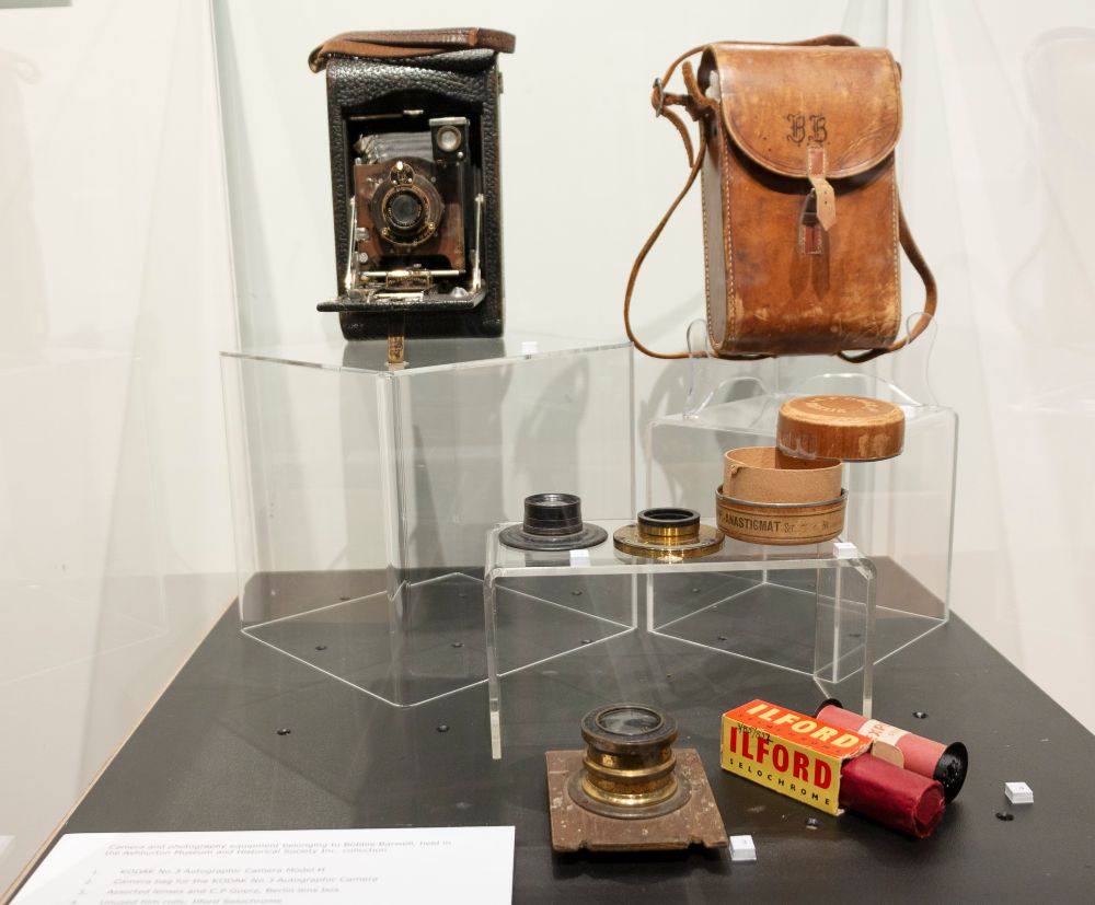 Bobbie Barwell’s No. 3 Autographic Kodak Model H, a folding bellows roll film camera, and accessories, including assorted lenses, unused rolls of film and a camera bag inscribed with the initials B.B., displayed in Bobbie Barwell: Capturing People and Places, Ashburton Art Gallery and Museum, 2022. Bobbie Barwell’s No. 3 Autographic Kodak Model H, a folding bellows roll film camera, and accessories, including assorted lenses, unused rolls of film and a camera bag inscribed with the initials B.B., displayed in Bobbie Barwell: Capturing People and Places, Ashburton Art Gallery and Museum, 2022.