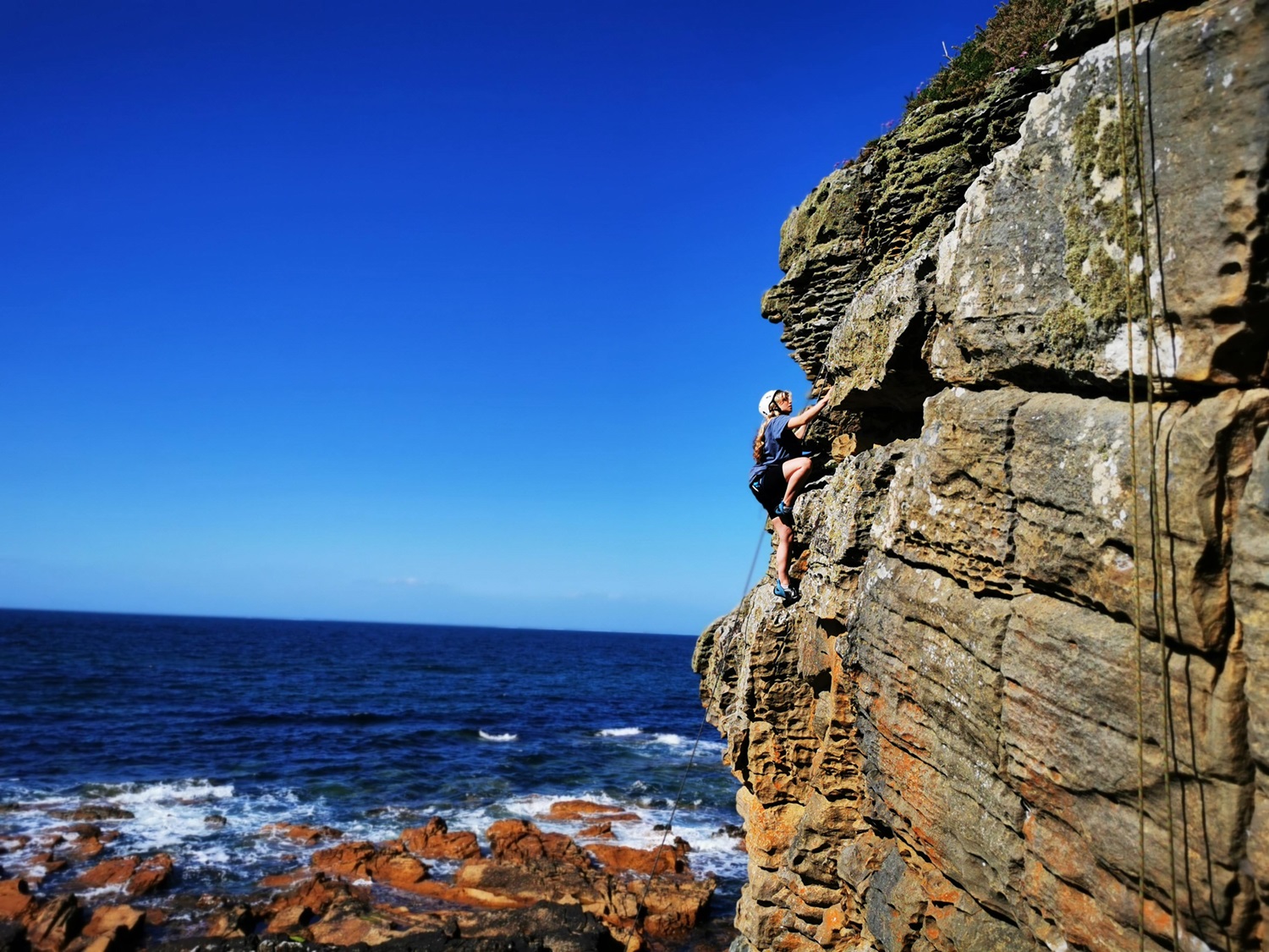 Deep blue sky with blue sea, and rocks in the foreground. A cliff juts out on the right with a woman stretching up to climb to the next hand and foot hold. Ropes can be seen hanging down the cliff face. It is a sunny day.