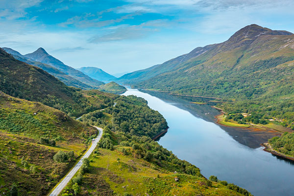 Picturesque view of a winding road down a glen with a loch reflecting the blue sky